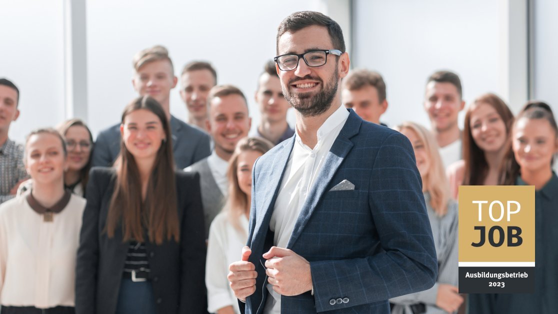 Lächelnder Mann im Sakko vor einer Gruppe junger Menschen, rechts das Siegel TOP JOB-Ausbildungsbetrieb 2023